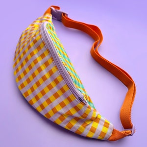 May include: A fanny pack with a yellow, orange, and purple gingham pattern. It has a purple zip and an orange strap. The background is a solid purple colour.