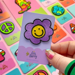 Op de afbeelding: Paarse geborduurde bloemenpatch met een gele smiley. De patch bevindt zich op een witte kaart met de tekst "by liz harry @lizharrydesign".