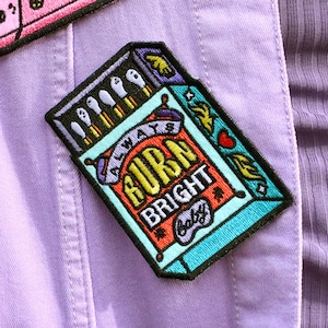 May include: A colourful embroidered patch featuring a matchbox design. The matchbox has a red and orange label that reads "Always Burn Bright Baby".