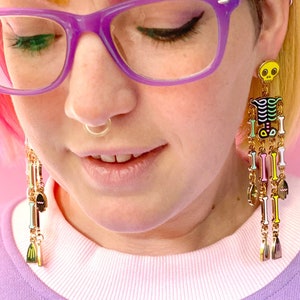 May include: Long, dangling earrings featuring a skeleton design with a yellow skull and colorful bones. The earrings have gold-colored chains and charms. The person in the image is wearing purple glasses and a light purple shirt.
