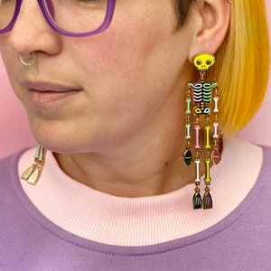 May include: A pair of long, colorful skeleton earrings. The earrings feature a yellow skull, a black torso, and dangling bone-shaped segments in various colors. The earrings are worn by a person wearing purple glasses and a purple and pink shirt.