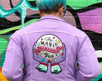 May include: A lavender denim jacket with an embroidered patch on the back. The patch features a pink and blue crystal ball with stars and the text "The Magic is Already in You".