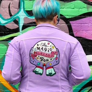 May include: A lavender denim jacket with an embroidered patch on the back. The patch features a pink and blue crystal ball with stars and the text "The Magic is Already in You".