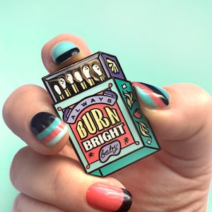 Op de afbeelding: Een turquoise blauwe emaille pin in de vorm van een luciferdoosje met de tekst "Always Burn Bright Baby" in witte en oranje letters. De pin heeft een afbeelding van lucifers met rode en gele vlammen.