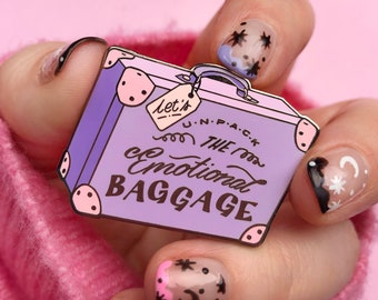Op de afbeelding: Paarse emaille pin in de vorm van een koffer met de tekst "Let's unpack the emotional baggage". De pin wordt vastgehouden door een hand met gelakte nagels.
