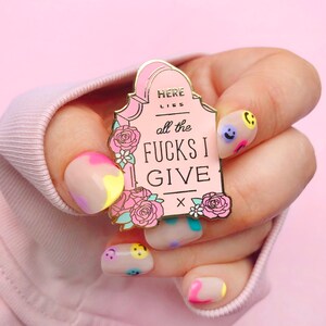 May include: A pink enamel pin shaped like a tombstone with the text "Here Lies All The Fucks I Give" and a small pink X at the bottom. The pin is surrounded by pink flowers and is being held by a hand with colorful nail polish.