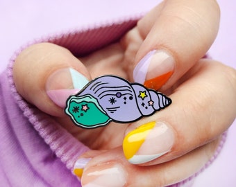 May include: A purple, green and yellow enamel pin shaped like a seashell with stars and a crescent moon. The pin is on a hand with colourful nail polish.