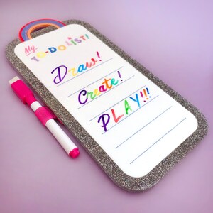 May include: A white dry-erase board with a rainbow-colored rainbow and the words "My To-Do List!" and "Draw! Create! PLAY!!!" in colorful lettering. A pink and white dry-erase marker is included. The board has a glittery silver border.