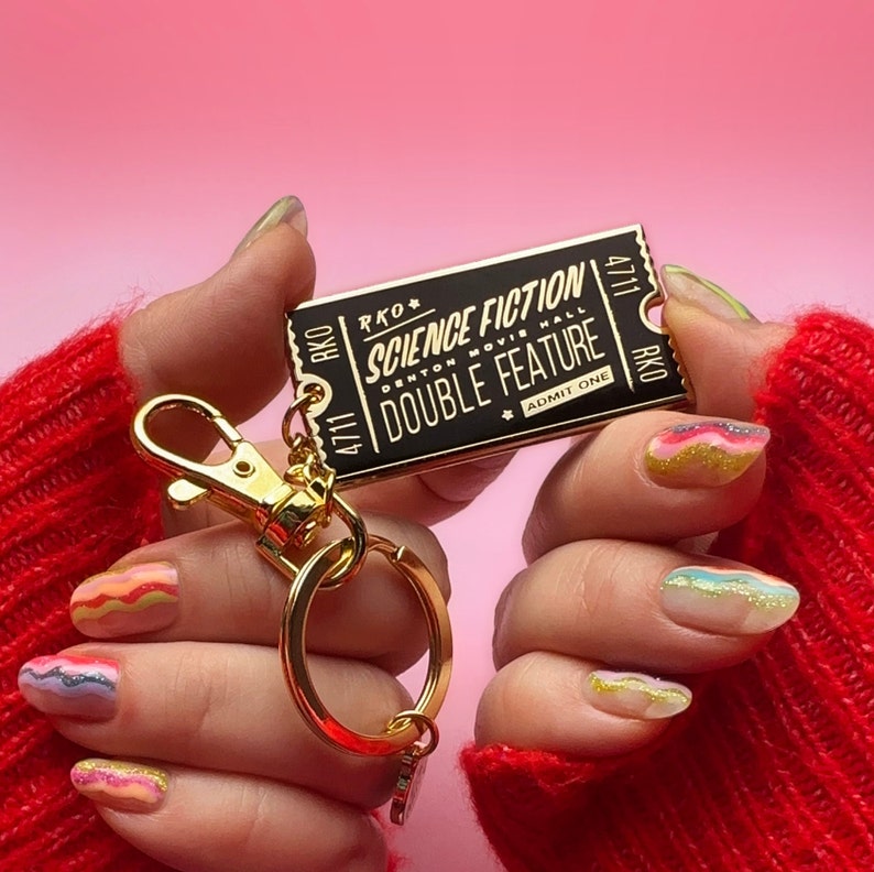 May include: Gold keyring with a black ticket-shaped charm that says "Science Fiction" and "Double Feature" with the words "RKO Denton Movie Hall" in smaller font. The keyring is held by hands with colourful nail polish.