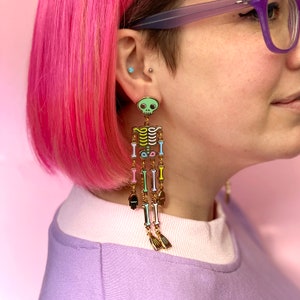 May include: A colorful skeleton earring with a light green skull and gold-colored bones. The earring features a series of colorful bones in shades of yellow, pink, blue, and green, with small hand and fin charms at the bottom. The earring is worn on the ear.