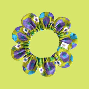 Op de afbeelding: Een groene bloemenkrans van stof met een paarse en blauwe bloemenprint. De krans is gemaakt van 10 bloemblaadjes en heeft een geplooide kern. De krans heeft een wit label met de tekst "Viva Flora" in zwart.