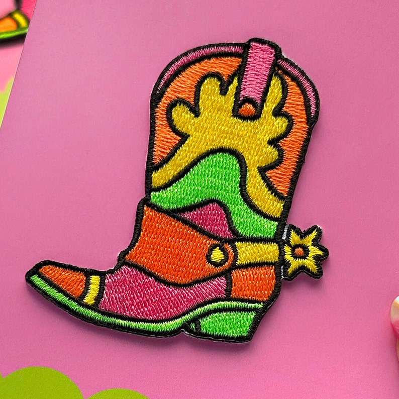 May include: A colorful embroidered patch of a cowboy boot with a pink, orange, green, and yellow design. The boot has a pointed toe and a spur on the heel.