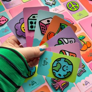 May include: A collection of colorful embroidered iron-on patches featuring various designs, including a dice, a flower, a cherry, a planet, and a unicorn. The patches are displayed on pink and white cards with the text "Liz Harry" and "@lizharrydesign".