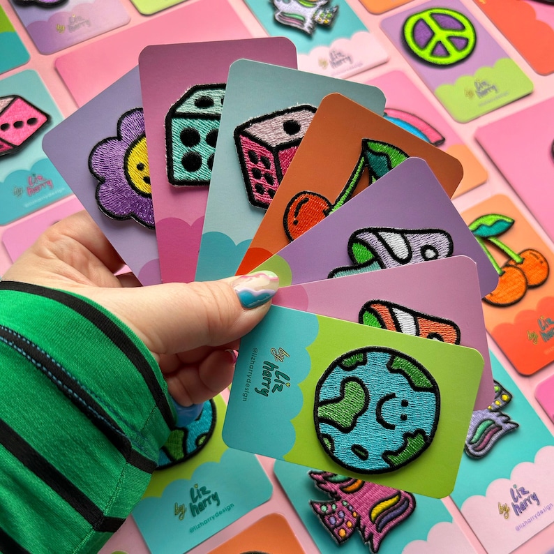 May include: A collection of colorful embroidered iron-on patches featuring various designs, including a dice, a flower, a cherry, a planet, and a unicorn. The patches are displayed on pink and white cards with the text "Liz Harry" and "@lizharrydesign".