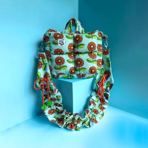 Może przedstawiać: Jasnoniebieska, pikowana torba typu tote z kwiatowym wzorem pomarańczowych kwiatów i zielonych liści. Torba ma pasujący pasek i małą metkę z napisem "HOT". Pasek jest ozdobiony pomarańczowymi sznurkami i zielonymi koralikami.