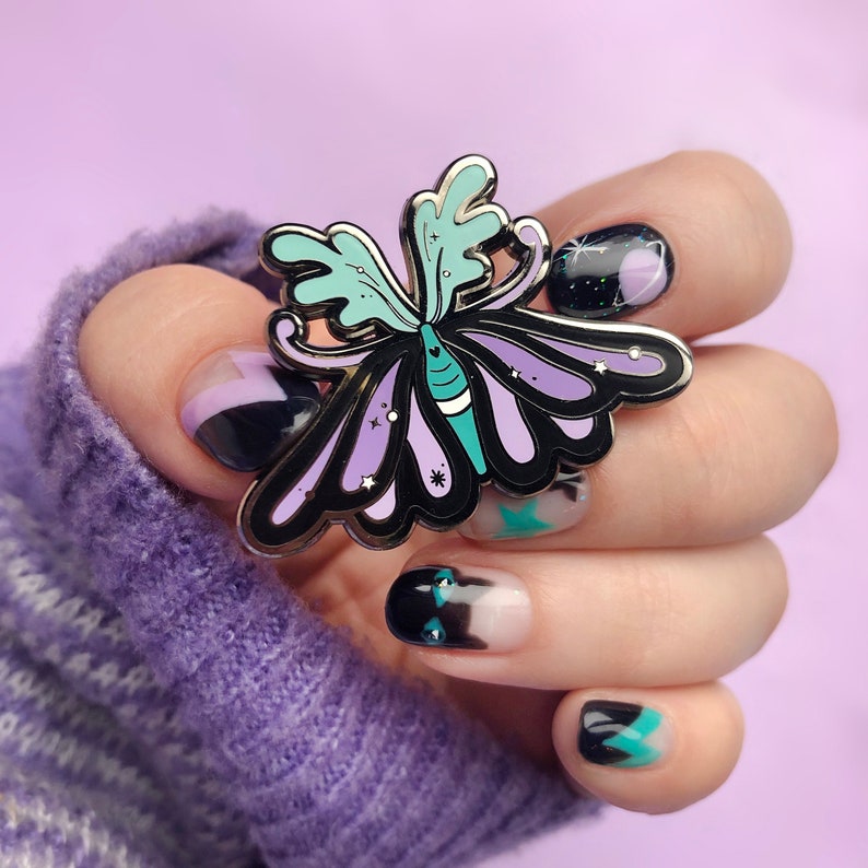 May include: A purple and teal butterfly enamel pin with a black outline and glitter accents. The pin is being held by a hand with black and white nail art.