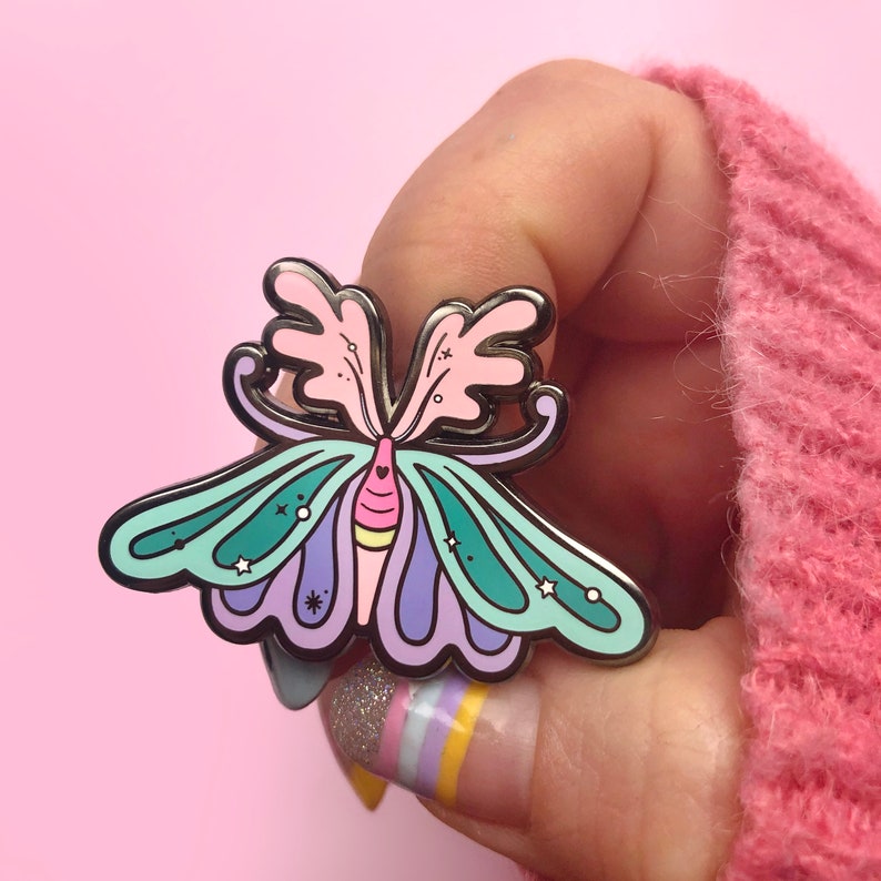 May include: A colorful enamel pin featuring a whimsical butterfly with pink, purple, green, and yellow accents. The butterfly has a heart-shaped design on its body and stars on its wings.