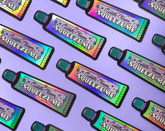 Può includere: Un motivo ripetuto di adesivi olografici a forma di tubetti di crema. Ogni tubetto ha un tappo verde e un'etichetta con la scritta "Cuddle Cream Squeeze Me & Apply Liberally", con stelle e cuori.