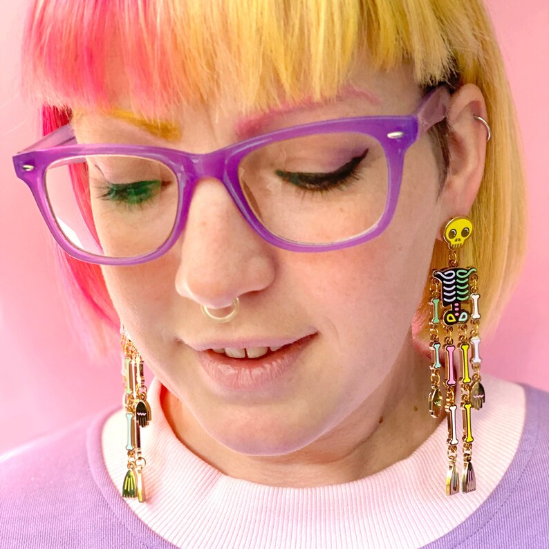 May include: Long, dangling earrings featuring a skeleton design with a yellow skull and colorful bones. The earrings have gold-colored chains and charms. The person in the image is wearing purple glasses and a light purple shirt.