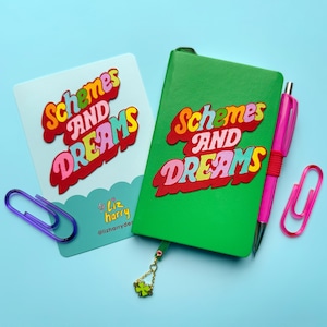 May include: A green journal and a light blue card with the words "Schemes AND DREAMS" in colorful, bubble letters. A pink pen, a pink paper clip, and a purple paper clip are also present. The journal has a charm with a four-leaf clover.
