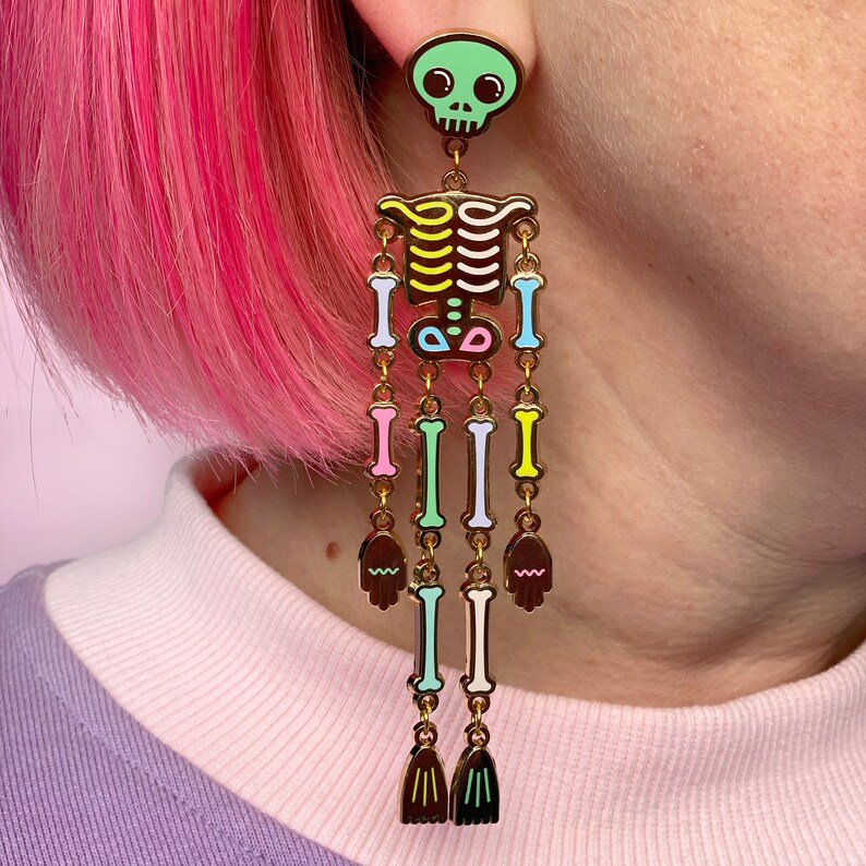 May include: A gold-colored skeleton-themed earring with a green skull top. The earring features a colorful rib cage and bones in pastel shades of pink, blue, and yellow. The earring has a dangling design with small hand and fringe details.