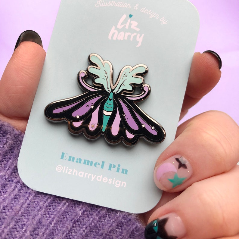 May include: A silver enamel pin featuring a purple and teal butterfly with black outlines and white stars. The pin is on a white card with the text "Illustration & design by Liz harry" and "Enamel Pin @lizharrydesign".