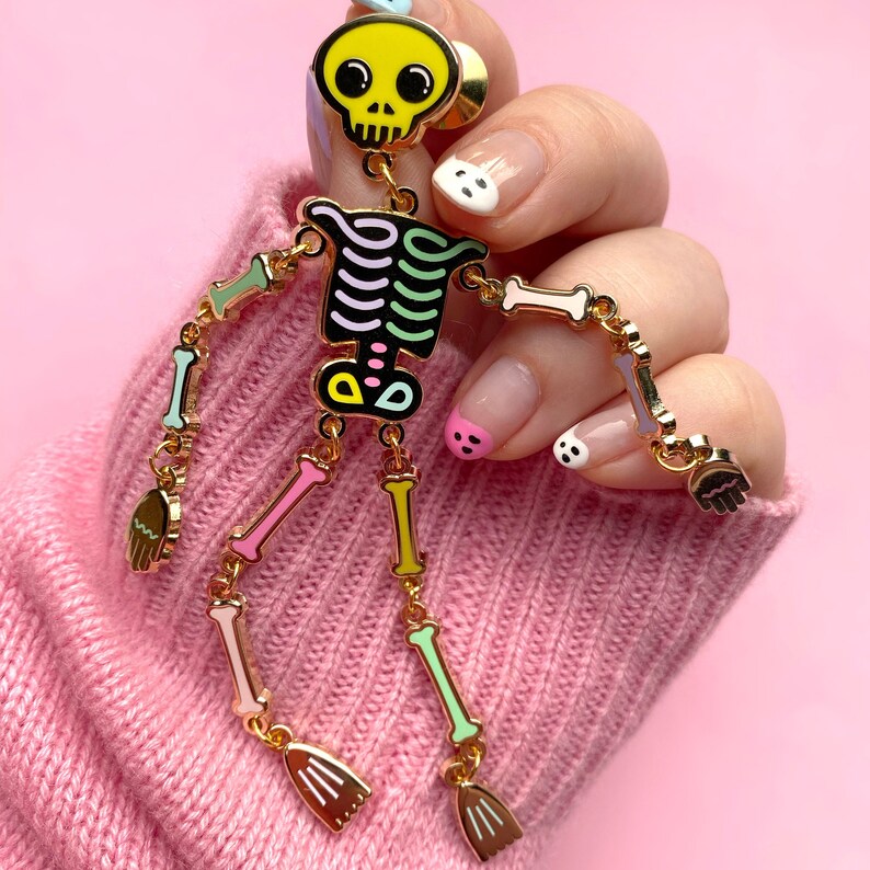 May include: A gold-colored skeleton pin with a yellow skull head and colorful bones. The skeleton's arms and legs are articulated, and it has small broom-shaped hands and feet. The pin is set against a pink background.