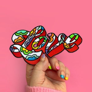 Puede incluir: Un parche bordado rojo con la palabra "LOVE" en letras coloridas y en espiral. Las letras están delineadas en blanco y rellenas con una variedad de colores, incluyendo rosa, amarillo, azul y verde.