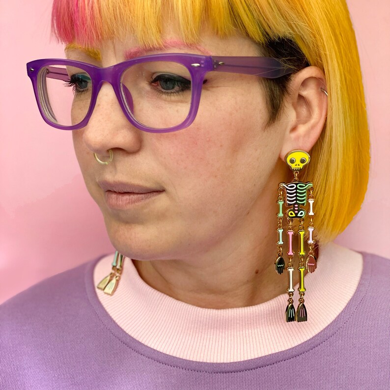 May include: A pair of long, colorful skeleton earrings. The earrings feature a yellow skull, a black torso, and dangling bone-shaped segments in various colors. The earrings are worn by a person wearing purple glasses and a purple and pink shirt.