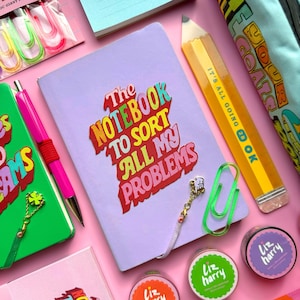 May include: A selection of stationery items on a pink surface. Features a lilac notebook with the text "The Notebook to Sort All My Problems", a yellow pencil-shaped ruler, and various colorful paper clips. Also includes pens, washi tape, and a green notebook.