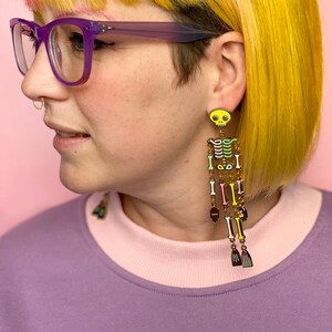 May include: Long, dangling skeleton earrings with a yellow skull top and colorful bone details. The earrings feature a gold chain and various colored bones, with small broom-shaped charms at the bottom. The earrings are worn by a person with yellow hair and purple glasses.