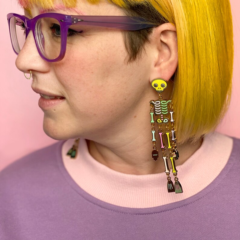May include: Long, dangling skeleton earrings with a yellow skull top and colorful bone details. The earrings feature a gold chain and various colored bones, with small broom-shaped charms at the bottom. The earrings are worn by a person with yellow hair and purple glasses.