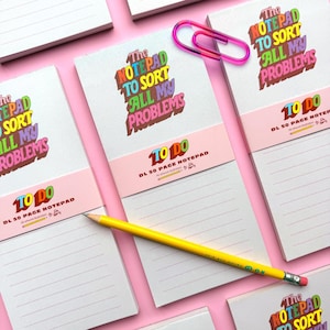May include: Several white lined notepads with the text "The Notepad to Sort All My Problems" in colourful lettering. A yellow pencil and a pink paperclip are also present. The notepads are labelled "To Do" and "DL 50 Page Notepad".