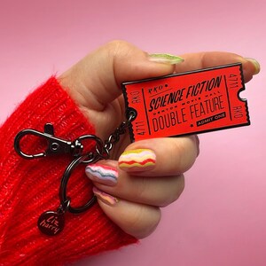 May include: A red metal keychain shaped like a movie ticket with the text "RKO Science Fiction Denton Movie Hall Double Feature Admit One" and the number 4711. The keychain is attached to a silver key ring with a small round charm that says "Liz harry".