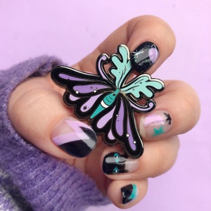 May include: A black metal butterfly pin with purple and teal wings. The pin has a sparkly finish and is shown on a hand with black and purple nail polish.