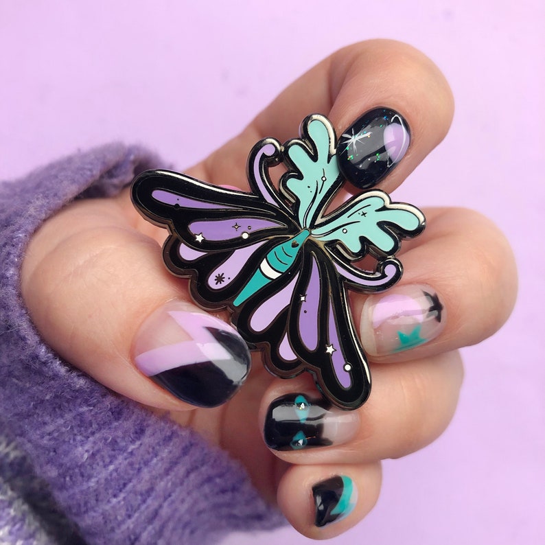 May include: A black metal butterfly pin with purple and teal wings. The pin has a sparkly finish and is shown on a hand with black and purple nail polish.