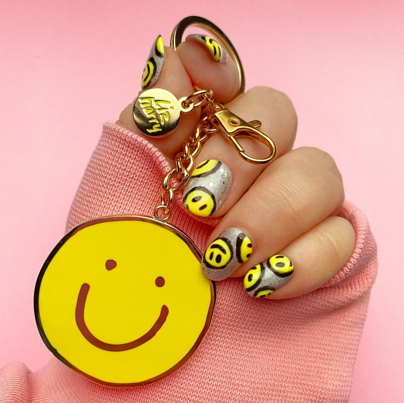 以下が含まれることがあります： 黄色いスマイリーフェイスのチャームと「Life Happy」と書かれた小さなチャームが付いたゴールドのキーチェーン。手には黄色いスマイリーフェイスのデザインのネイルポリッシュが塗られています。
