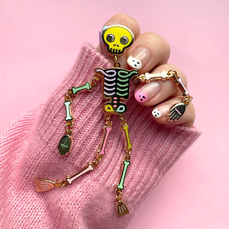 May include: A whimsical skeleton pin with a yellow skull, black accents, and colorful ribcage and bones. The pin is gold-toned and has articulated arms and legs, with small hand and foot charms. The pin is displayed against a pink background.