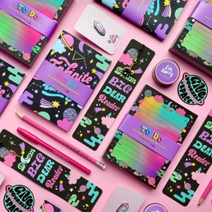 Puede incluir: Una colección de artículos de papelería coloridos con un tema espacial. Los artículos incluyen cuadernos, listas de tareas, marcapáginas y pegatinas. Los diseños presentan planetas, estrellas y nubes en una paleta de colores arcoíris.