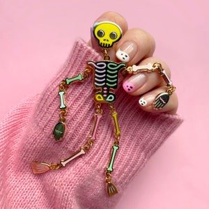 May include: A colorful skeleton pin with a yellow skull, black and green rib cage, and pastel-colored bones. The pin is gold-toned and has articulated arms and legs, with small broom-shaped hands. The pin is displayed against a pink background.