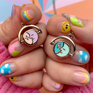 May include: Two gold necklaces with a spinning charm featuring a blue and pink moon with a star and a closed eye. The necklaces are worn on two hands with colourful nail polish.