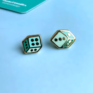 May include: A pair of gold-tone earrings shaped like dice. One die is white with black dots and the other is light blue with black dots.