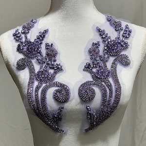 May include: A pair of lilac-colored beaded applique embellishments with a floral design. The embellishments are made of rhinestones and beads on a sheer fabric base.