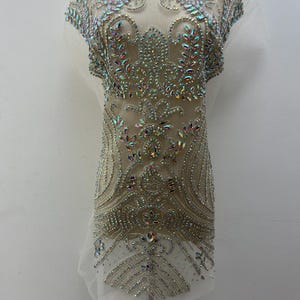 May include: A white mesh fabric with a large, intricate design of iridescent rhinestones and beads. The design features a floral pattern with a central motif that resembles a heart. The fabric is displayed on a mannequin.