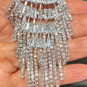 May include: A silver-toned, crystal-encrusted, teardrop-shaped dangle earring with a cascading design. The earring features multiple rows of baguette-shaped crystals and smaller round crystals.