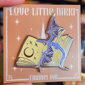 May include: An enamel pin featuring a blue dragon with purple wings perched on an open book with yellow pages. The book displays a crescent moon and star symbols. The pin is attached to a square tan backing card with the text Love Little Nikki Enamel Pin.