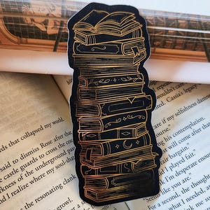 Peut inclure: Autocollant noir représentant une pile de livres avec des détails dorés. Les livres sont de tailles variées et présentent des éléments décoratifs. L'autocollant est placé sur un fond de pages de livre ouvertes.