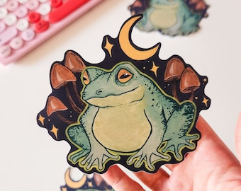 Puede incluir: Una pegatina con una rana verde sentada en un lecho de champiñones marrones con una luna creciente amarilla y estrellas en el fondo.