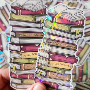 Könnte beinhalten: Aufkleberbogen mit Stapeln von Büchern in verschiedenen Farben, darunter Burgunderrot, Grün und Gold. Die Aufkleber haben einen holografischen Effekt, wobei einige Bücher geöffnet sind. Die Aufkleber werden von einer Hand gehalten.