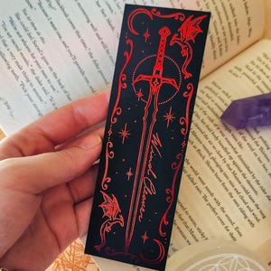 May include: Black bookmark with a red sword design, the words "Wind Cleaver", and dragon illustrations. The bookmark is held over an open book.